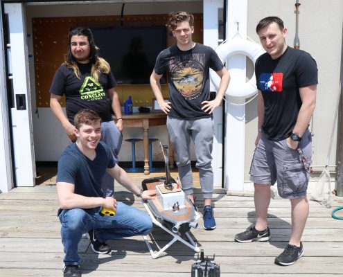 Four male students stand on the dock at the MIT Sailing Pavilion with a surface vehicle resembling a miniature boat that they developed