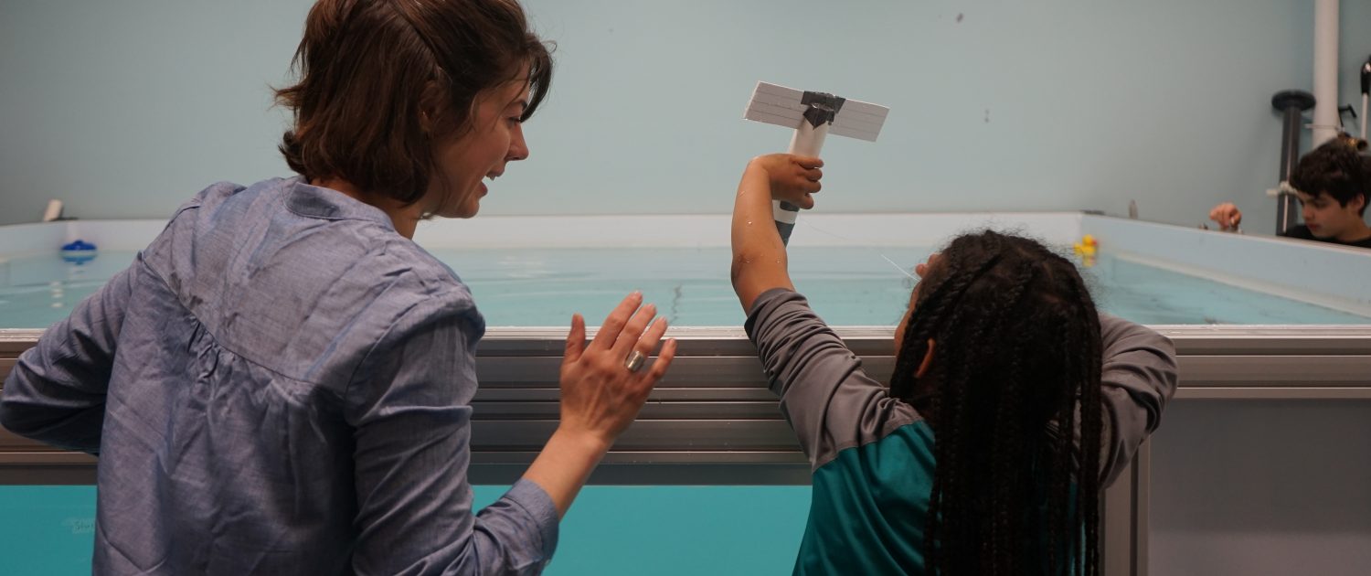 A woman and a child from behind about to drop a cardboard model into a large tank of water.